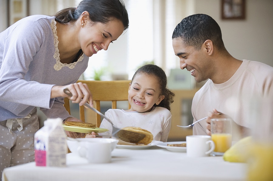 Family eating breakfast together in the morning with the mom serving the child in the middle a pancake as the mom, child and father smile