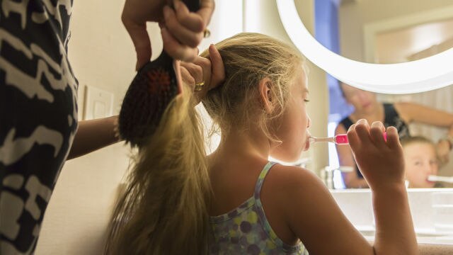 Girl brushing her teeth while her hair is being combed