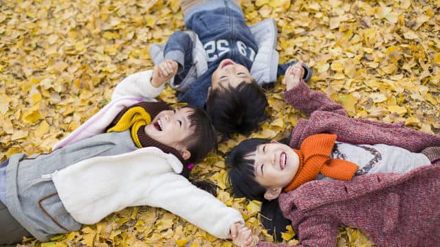 three kids smiling while laying down on the ground while looking up, and holding hands
