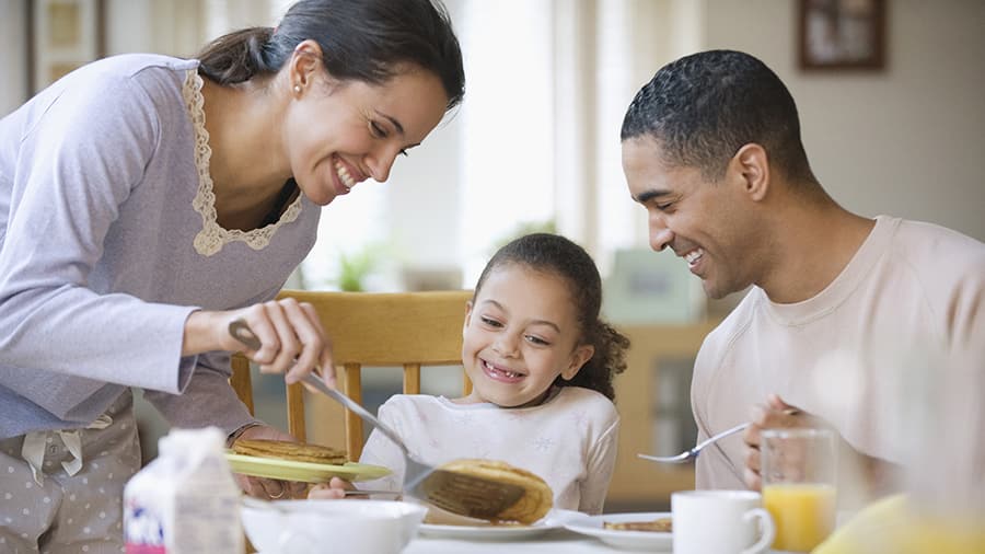 Family eating breakfast together in the morning with the mom serving the child in the middle a pancake as the mom, child and father smile